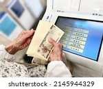 Small photo of Hands of an elderly woman counting bills in front of an ATM machine. Japanese means Deposit, withdrawal, balance inquiry, transfer, passbook entry, various registrations, foreign currency deposits.