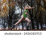 A dancer mid-leap, wearing a green bodysuit and fishnet tights, against a backdrop of fall foliage at dusk.