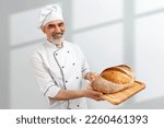 Small photo of Chef-cooker in chef's hat and jacket working in bakery, holding French bread board with bread. Senior professional baker man wearing chef's outfit. Character kitchener, pastry chef for advertising.