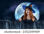 Halloween Witch peeking out behind a fence or wooden wall. Female wizard fairy character. All Saints' Day. Fantasy gothic red-haired sorceress girl dressed in black carnival costume. Enchantress woman