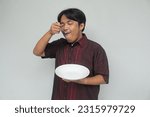Small photo of Young Asian man holding empty plate and shoving spoon to mouth with happy and full expression.