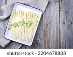 Traditional steamed white asparagus with sauce hollandaise and herbs as top view in an enamel tray on a wooden board with copy space right