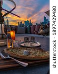 Small photo of Manakish with shisha spoon wooden plank and hot mint tea being poured out with cityscape in background whilst sunset is taking place with orange glow. Focus is centered on tea glass. Portrait mode
