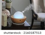 Small photo of An oil diffuser that is on and producing a thick, steady stream of mist. It's diffusing essential oils while playing music and changing colors. The round remote is next to the machine.