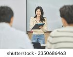 Small photo of An Asian female student or professor is wearing a mask and standing in front of a lecture hall at a university in Korea, giving a presentation and lecture. Male students are sitting in front of her