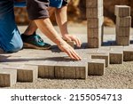 Young man laying gray concrete paving slabs in house courtyard on gravel foundation base. Master lays paving stones. Garden brick pathway paving by professional paver worker. Repairing sidewalk.