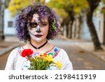 front view portrait of serious young woman with curlers holding flowers and with artistic makeup representing La Calavera Catrina, standing outdoors looking at camera with copy space.