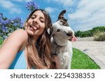 Selfie portrait of young Colombian Latina woman, with her border collie dog, in the park sticking out her tongue and looking at the camera, with the sky and trees in the background, phone perspective.