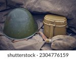 Military flask in a case with a military helmet and mess kit in blur. Close up WW2 army ammunition. The concept of the memory of the fallen warriors. Reconstruction in the trench. Selective focus.