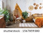 Elegant and quiet bohemian room with cozy interior, wicker chair, pillows, cushions, green plants in flower pot, bed and rug on wooden floor