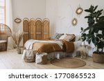 Comfortable apartment in bohemian style interior with hygge bedroom, pillow and bedspread on bed, bamboo dressing screen, home decor, dry plants in vase, wicker basket, houseplant on floor