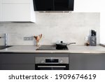 Kitchen with built in appliance, electric cooker hood, stove, oven and sink. Frying pan on glass ceramic surface at marble countertop close to wooden spoons and knives, under white cupboards