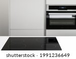 White countertop with glossy built in ceramic black induction stove, big white cupboard with built-in electrical oven in empty kitchen with nobody in sight, blurred background