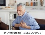 Concept of health and cardiovascular problems. Senior man with cardiology disease feeling sharp heart pain, holding hands on chest, sitting on armchair at home. Pensioner need emergency medical care