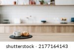 Croissant and coffee on kitchen countertop, against blurred minimalist interior with modern furniture. Selective focus at homemade pastry and tea drink in cup on wooden table, copy space, web banner