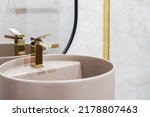 Close up shot of golden shiny water tap over washbasin. Pink wash basin with faucet in classic style in light bathroom against copy space on tiled wall. Concept s of morning routine, hygiene at home