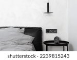Small photo of detail in bedroom interior, pillow and duvet in cotton case on bed with black leather headboard, alarm clock on metal coffee table and electrical socket with switch on white wall