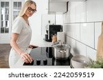 female housewife turn on knob switch at control panel of electrical glass ceramic hob to make dinner in saucepan at kitchen with modern interior