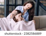 Sick child girl sitting on couch covered with blanket blowing nose, sneezing in tissues spending day at home with mother. Supportive mom hugging her ill daughter with flu symptoms