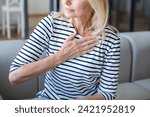 Small photo of Cropped shot of mature woman with sudden cardiac pain hold hand on chest and sitting on couch alone. worried female feeling discomfort and tachycardia and heart attack symptom. problems with health