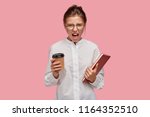 Small photo of Photo of petulant vexed college student looks angrily at camera, holds hot coffee in disposable cup, red book, feels crazy after constant preparation to exams, poses against pink background.