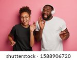 Small photo of Positive Afro woman and man sing new song and dance with pleasure during disco party, listen favourite music in headphones, excited with successful news, move against promotional pink background