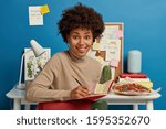 Small photo of Positive busy worker with Afro hairstyle makes notes in notepad, wears turtleneck, concentrated on coursework, writes new chapter of book, notes some records, smiles broadly, eats pizza at desktop