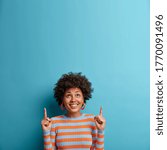 Small photo of Cheerful African American woman looks up and points upwards with broad smile, recommends awesome product, suggests click banner, dressed casually, isolated on blue background shows advertisement promo