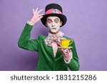 Small photo of Time for having tea. Aristocratic gentleman with bright makeup has image of fictional character holds cup of drink wears big hat has wondered expression poses over purple wall. Halloween celebration