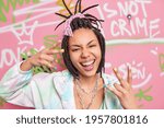 Small photo of Cool hipster girl makes rap gesture feels crazy and happy smiles broadly has cheeky expression dressed in stylish clothes poses near graffiti wall. Millennial generation art youth lifestyle concept