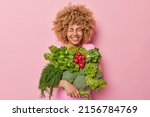 Small photo of Cheerful woman with curly hair carries different fresh vegetables keeps eyes closed smiles broadly isolated over pink background. Healthy organic food and spring harvest concept. Garden produce