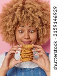 Small photo of Vertical shot of surprised curly haired young pretty woman holds pile of appetizing chocolate chip cookies feels temptation to eat sweet food poses indoor. Yummy harmful food and calories concept.