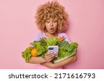 Small photo of Photo of shocked curly haired woman uses mobile phone for buying products online carries green fresh vegetables cannot believe own eyes poses against pink studio background. Grocery concept.