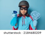 Photo of serious woman wears black protective helmet eyemask and rubber gloves dressed in superhero costume holds spray detergent and rag going to wash everything dirty in room isolated over blue