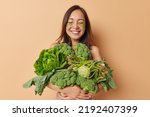 Small photo of Pleased smiling Asian woman applies beauty patches under eyes to reduce bags poses with variety of fresh green vegetables full of vitamins eats asparagus kohlrabi and broccoli isolated over beige