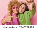 Small photo of Best female friends have fun make frame gesture with fingers smile toothily wear casual jumpers foolish around isolated over pink background. Happy biracial women measure angle picture something