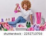 Happy curly haired woman picks up new high heeled sandals likes buying different shoes dressed in casual white t shirt and jeans poses on floor chooses best footwear to wear on special occasion