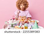 Small photo of Indoor shot of curly woman points at you with reproach feels sad poses near festive table with desserts cone hats celebrates birthday isolated over pink background. Festive event celebration concept