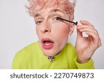 Small photo of Close up portrait of transgender guy puts on makeup. Young gay man applies mascara on eyelashes keeps mouth opened cares about his appearance wears green jumper isolated over white background