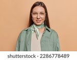 Small photo of Portrait of good looking young woman with dark straight hair looks directly at camera wears green shirt and kerchief around neck big spectacles isolated over brown background ready for work.