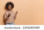 Small photo of Pretty young Afro American woman with curly bushy hair hods mobile phone texts and checks her newsfeed dressed in casual hoodie and jeans presents some information isolated over brown background