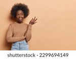 Small photo of Confused dark skinned woman shrugs shoulders looks unaware raises arm in bewilderment asks so what dressed in casual jumper jeans isolated over brown background copy space for your advertising concept