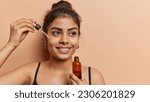 Small photo of Domestic skin care and anti wrinkle routine. Pleased dark haired woman applies facial serum with dropper smiles toohthily focused aside dressed in t shirt isolated over brown background copy space