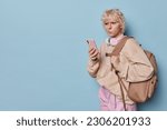 Small photo of Horizontal shot of displeased millennial girl concentrated at screen of smartphone feels unhappy carries big backpack surfs social networks checks newsfeed dressed casually poses over blue background