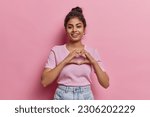 Small photo of Portrait of happy Indian woman with hair bun shows finger heart gesture sends love to someone smiles pleasantly wears casual clothes isolated on pink background expresses sympathy feels passionate