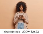 Small photo of Horizontal shot of dark skinned curly woman holds mobile phone in hands checks newsfeed and sends text messages wears shirt jeans poses against brown background. Modern technologies concept.