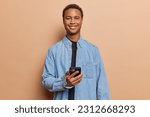 Small photo of Confident young dark skinned man dressed in stylish denim shirt poses with mobile phone in hand checks newsfeed has satisfied expression isolated on brown background. Technology and lifestyle concept