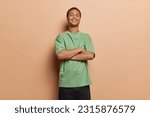 Isolated studio shot of dark skinned young man stands self confident keeps arms folded dressed in casual green t shirt and black trousers feels proud of himself poses against brown background.