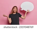 Small photo of Clueless hesitant European woman holds empty speech bubble in her hand embraces opportunity to fill void with unspoken sentiments shrugs shoulders wears black t shirt isolated over pink background