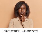 People emotions concept. Studio photo of young pretty thoughtful African american woman standing in centre isolated on beige background keeping right hand raised under chin looking straight at camera
